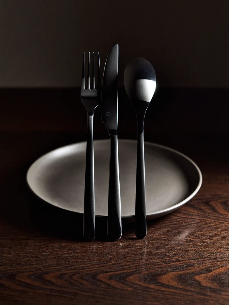Couverts noirs mats posés sur une assiette en céramique grise, sur une table en chêne sombre.