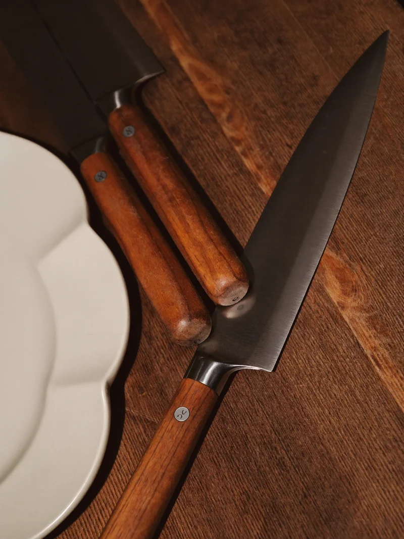 Trois couteaux à steak à manche en bois sur une planche de noyer, près d'une assiette blanche.