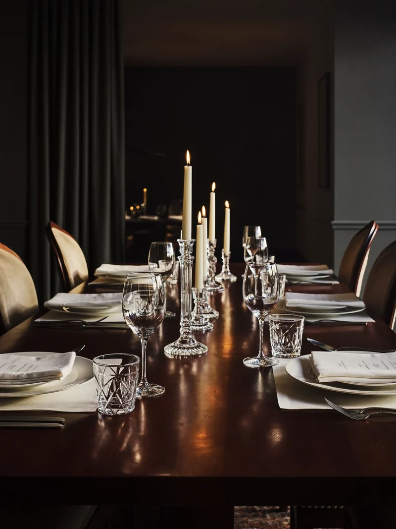 Longue table dressée pour six, avec couverts, verres sobres et bougies dans une salle à manger tamisée.